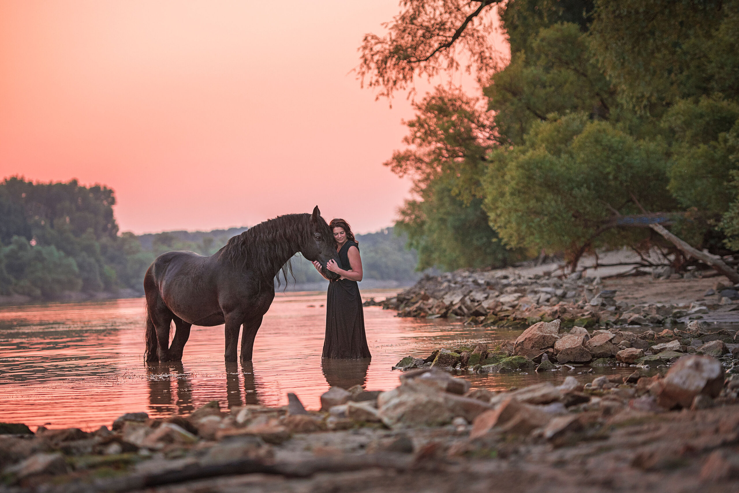 Eine Frau in einem schwarzen Kleid steht neben einem schwarzen Pferd im Wasser.