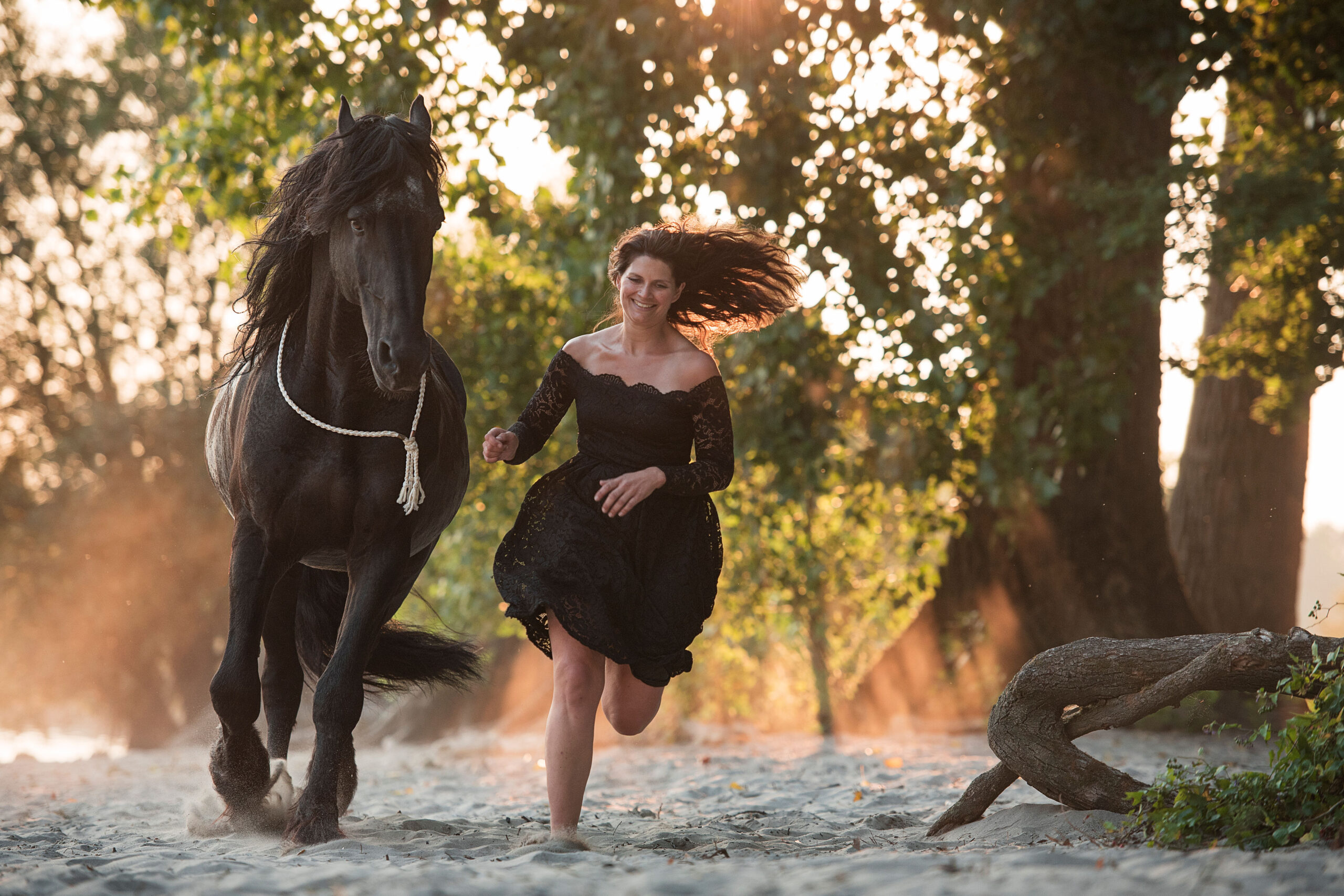 Eine Frau in einem schwarzen Kleid läuft neben einem schwarzen Pferd auf Sand.