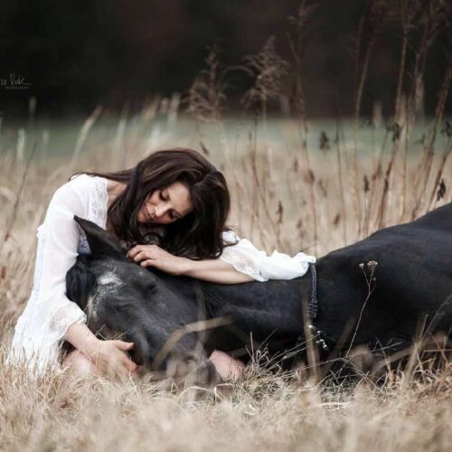 Eine Frau im weißen Kleid sitzt in einem Feld, auf ihrem Schoß liegt der Kopf eines Rappen.